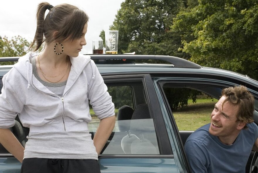 A young woman leaning on the passenger door of a car looking down at the male driver of the car who is sitting in the car and looking up at her.