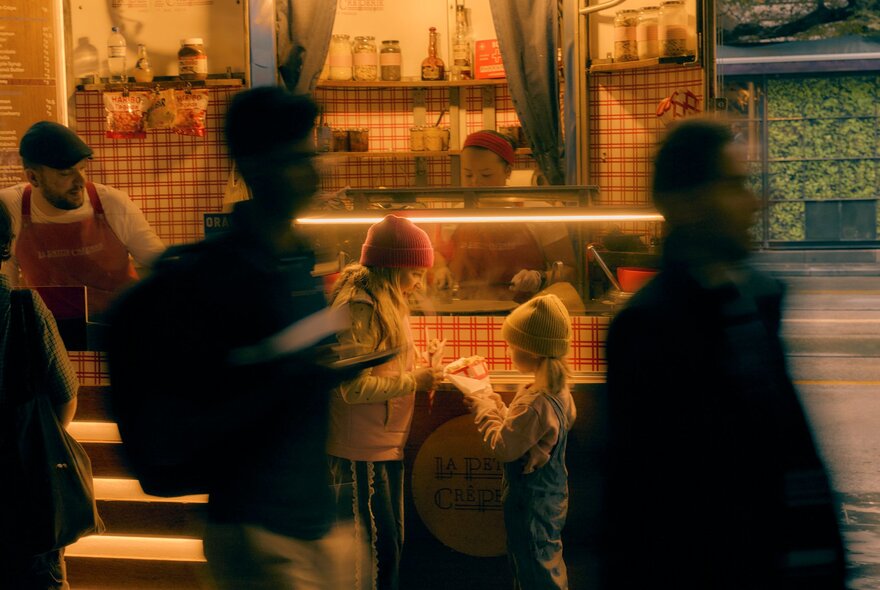 A nighttime street scene featuring a warmly lit food van selling crepes and two children wearing beanies eating crepes in front of it, blurred city commuters walking past in the foreground.