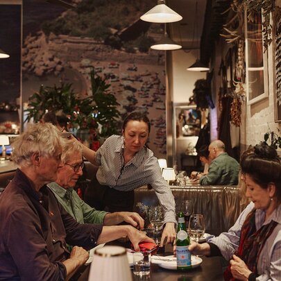 An indoor restaurant scene showing a group of people sitting at a table with a waiter placing plates of food in front of them.