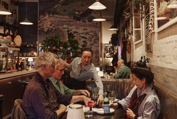 An indoor restaurant scene showing a group of people sitting at a table with a waiter placing plates of food in front of them.