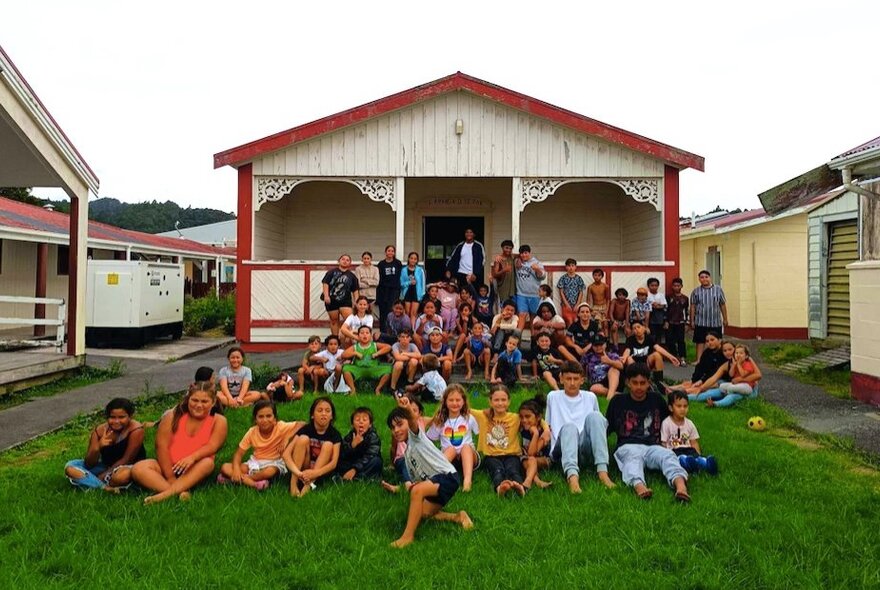 A group of people sitting on a lawn in front of wooden buildings.
