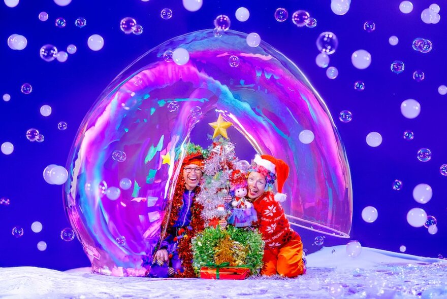 Two performers dressed in Christmas outfits inside a large pink bubble with a Christmas tree surrounded by bubbles.