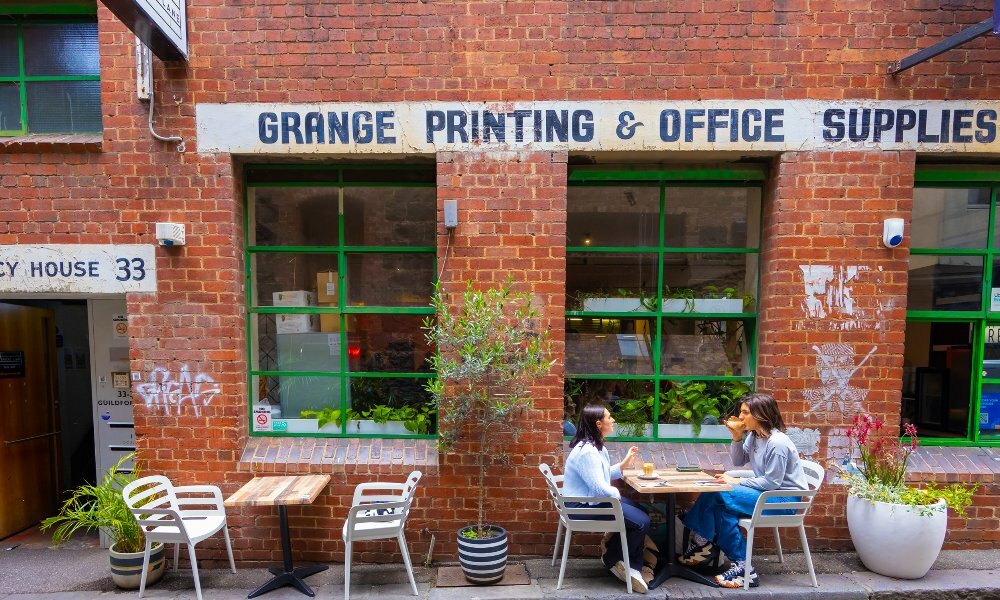 Two friends are sitting in front of a cafe with a brick exterior drinking coffee