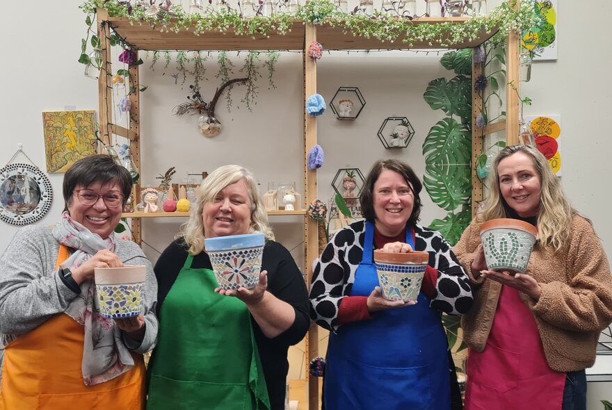 Workshop class participants smiling in a studio setting, holding up mosaic pots they have made.