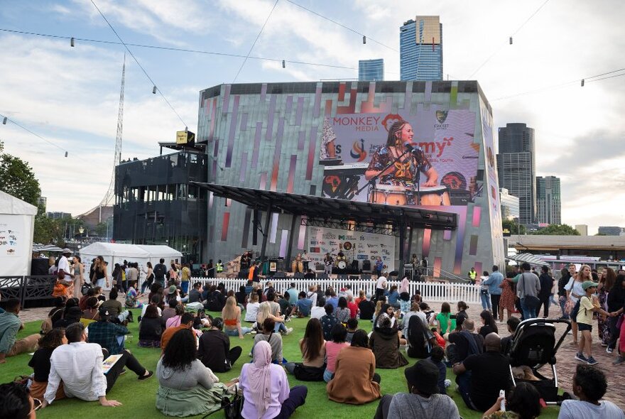 Crowd seated and seen from behind, watching performance on large screen, outdoors.