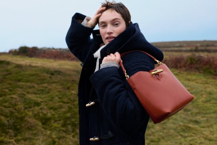 Model wearing a duffel coat on a windswept moor, holding a brown handbag.