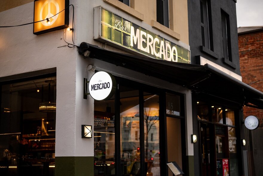 The exterior of Bar Mercado with a small illuminated round sign on the corner. 