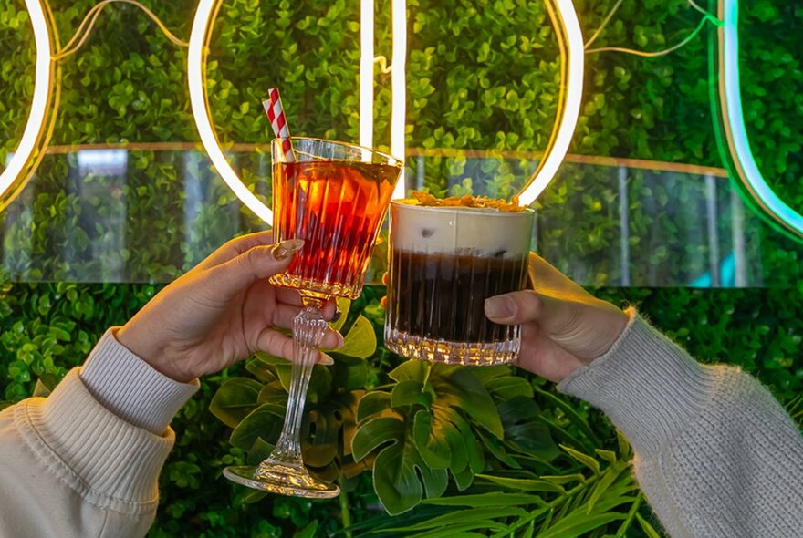 Two people are doing a cheers in front of a green leafy wall