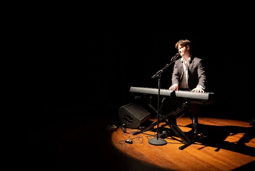 Performer seated on a spotlit stage and playing a keyboard while singing into a microphone.