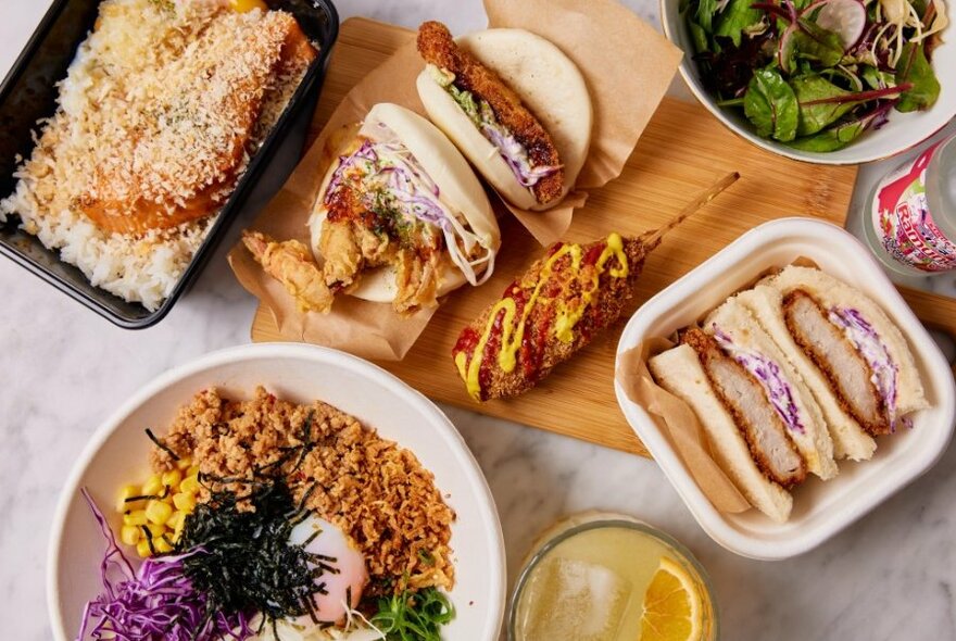 A table set with Japanese dishes including katsu sando, bao buns and rice bowls.