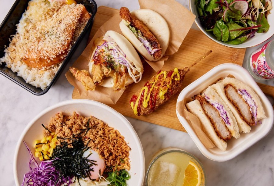 A table set with Japanese dishes including katsu sando, bao buns and rice bowls.