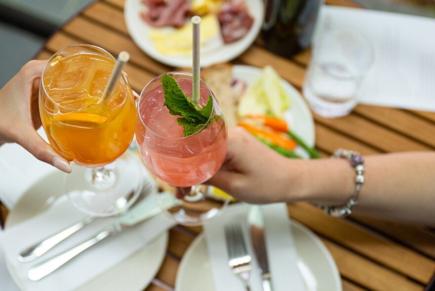 Two hands, each holding a summer cocktail, clinking glasses over a wooden table set with plates and cutlery.