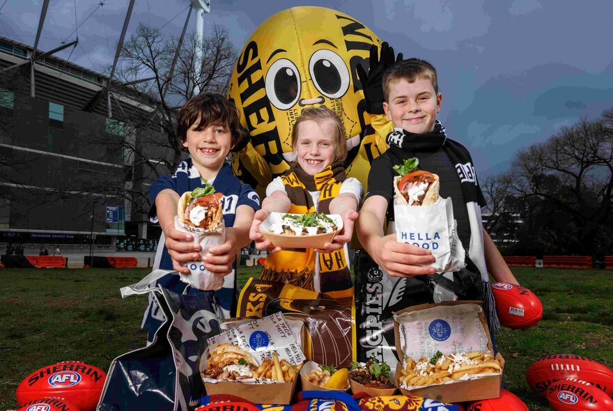 Three children wearing AFL football merchandise, holding out food, standing in front of a life size yellow Sherrin football at Yarra Park outside the MCG.