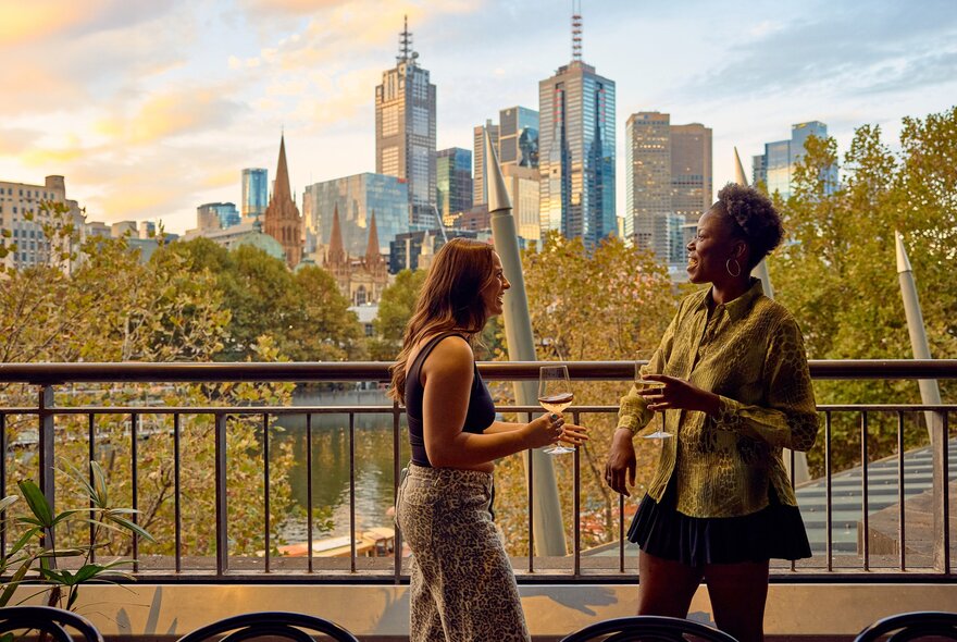 Two friends are drinking wine on a balcony over looking Melbourne's skyline and the Yarra river