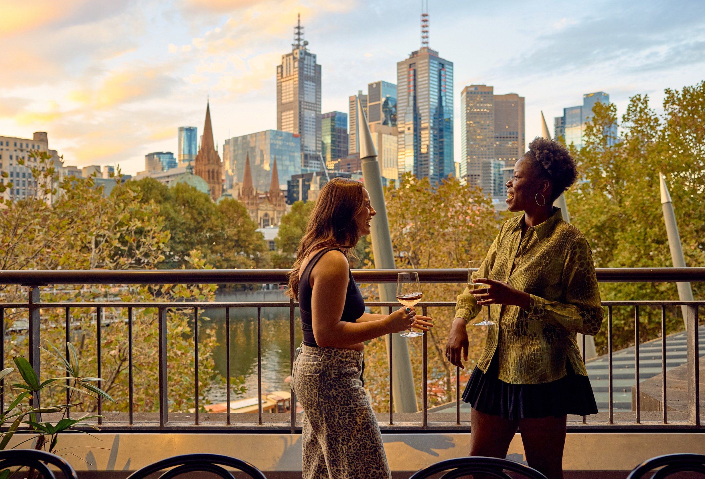 Two friends are drinking wine on a balcony over looking Melbourne's skyline and the Yarra river