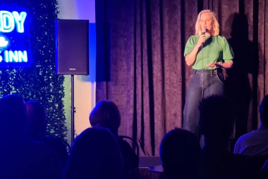 Female stand-up comedian talking into a microphone and standing on a stage, partial neon signage visible on the left of the image, seated audience in front of stage.