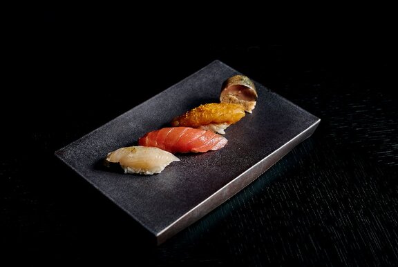 A black serving plate with four unique pieces of sushi, including kingfish, salmon, prawn and eel.