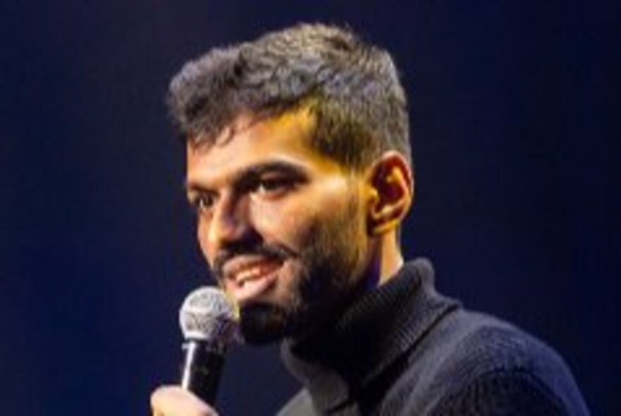 A comedian wearing a turtle neck jumper performing and holding a microphone.