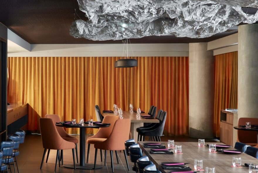 A restaurant interior with orange pleated curtains behind tables set for dinner.