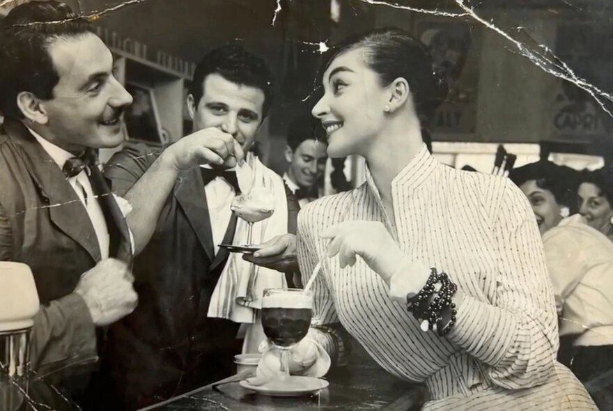 Vintage cafe image of woman with cocktail and Italian waiters wearing jackets and bow ties.