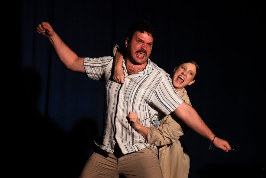 Two performers on stage, one standing behind a person and with their arms wrapped around them, both singing.