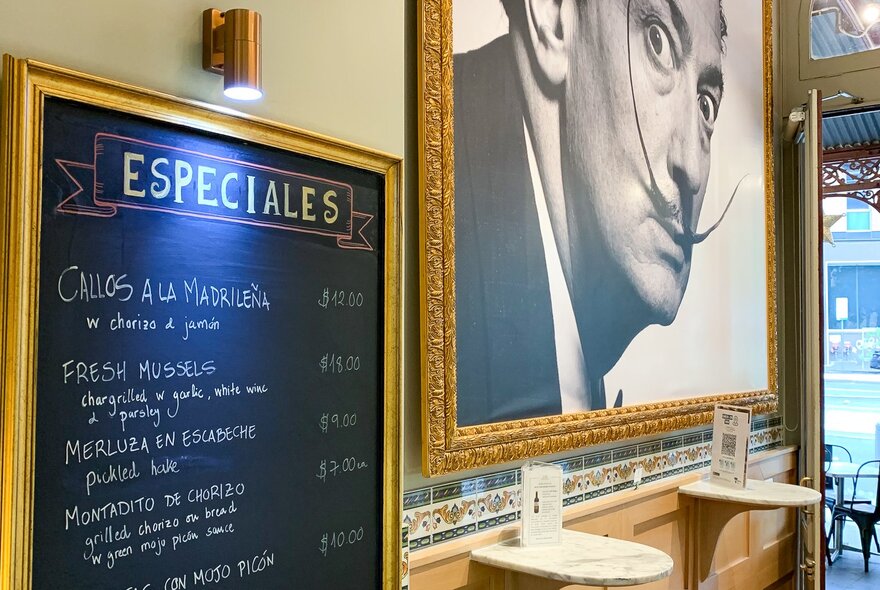 A menu written on a chalkboard next to a large photograph of Salvador Dai. 