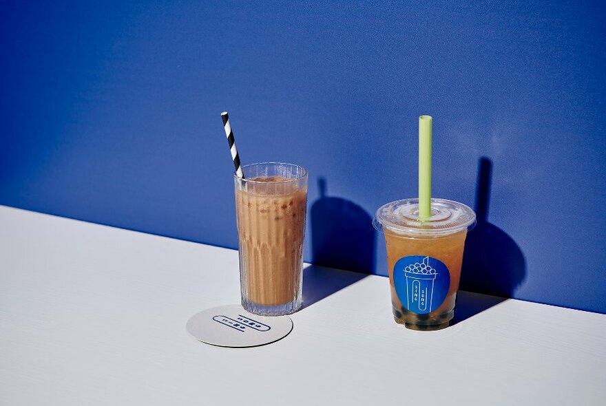 Two bubble tea drinks, one in a glass and the other in a plastic takeaway container, both with straws.