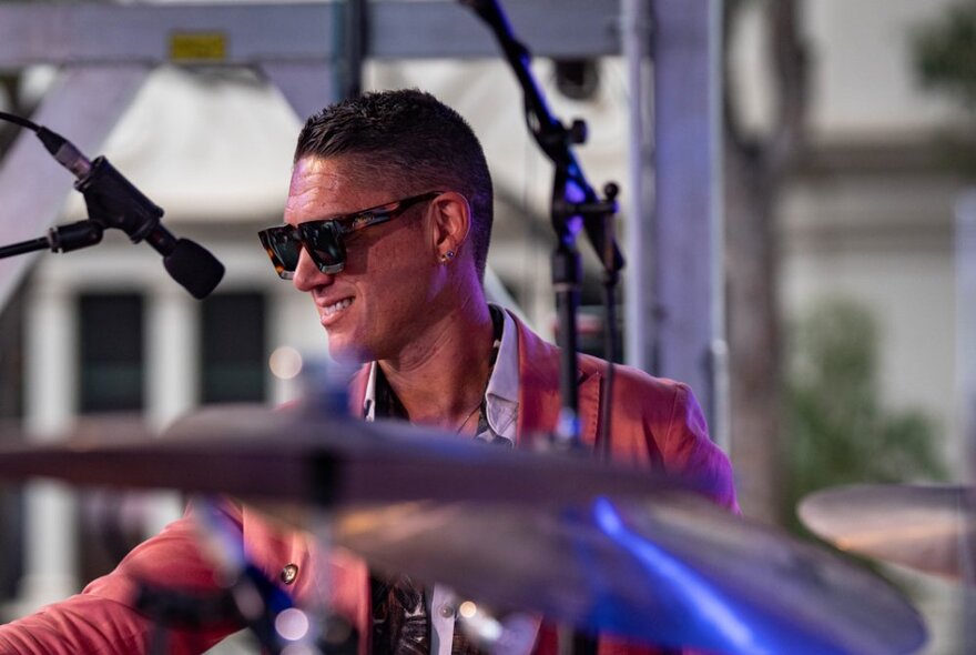 A smiling man in sunglasses and a salmon blazer with an out-of-focus drum kit in the foreground. 