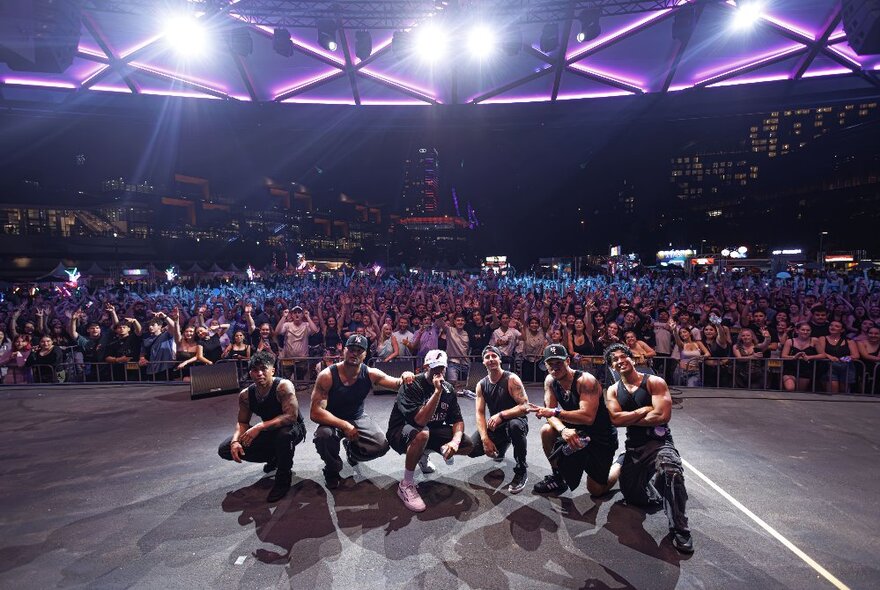 A dance troupe squat on stage with their back to a large audience, purple lights overhead. 
