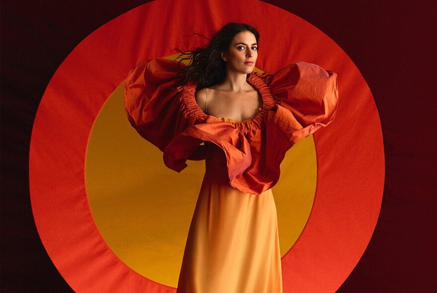 Singer Anna Setton in a dress with a large orange frilled collar against a background showing an orange and yellow sun.