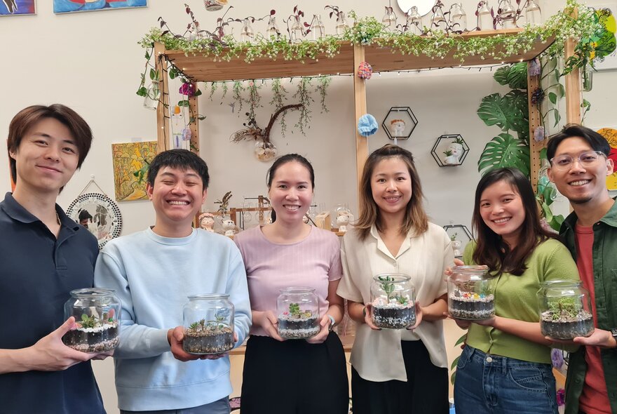 A group of smiling participants in a studio workshop setting, holding glass-domed terrariums.