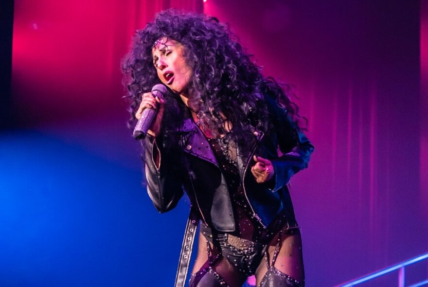 A tribute performer dressed as Cher singing soulfully with a blue and red lit background. 