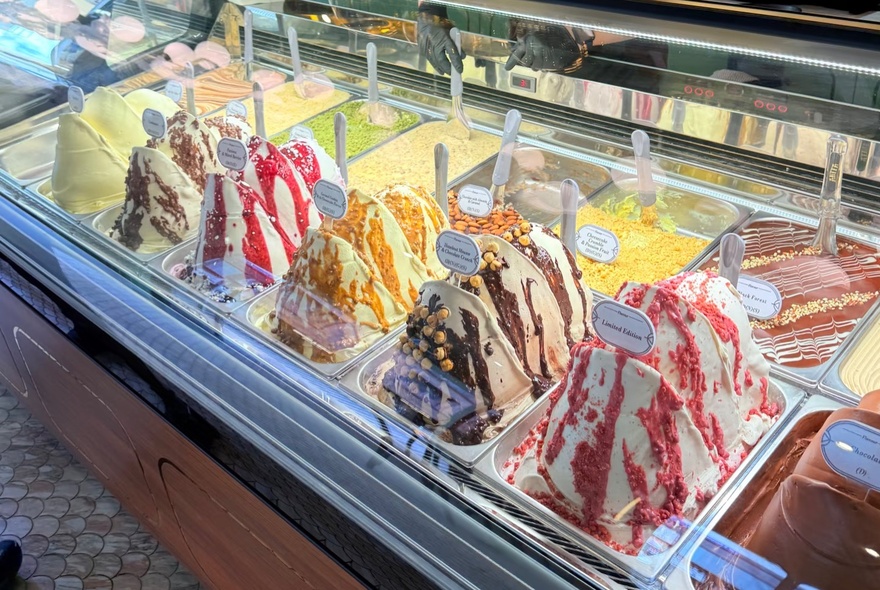 Multi-coloured ice cream and gelato in metal tubs inside a refrigerated glass display cabinet.