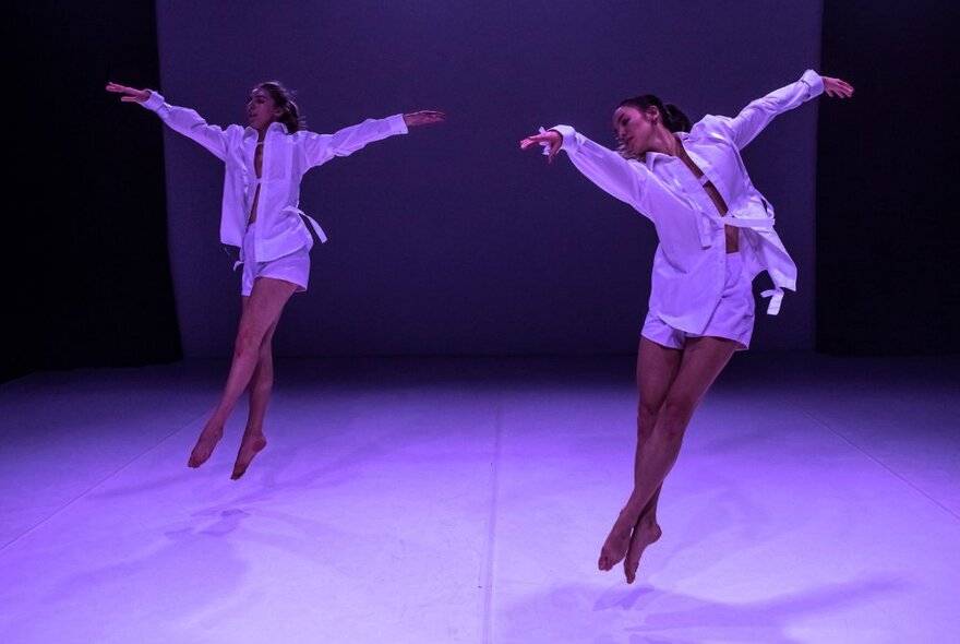 Two dancers with their arms outstretched and jumping in mid air on a stage.