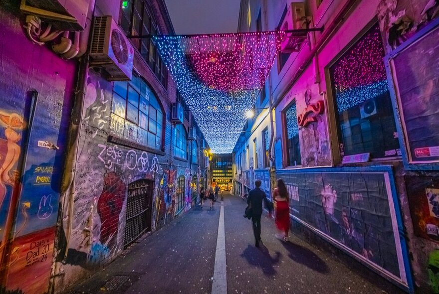 People walking down a laneway with street art and a row of colourful lights overhead.