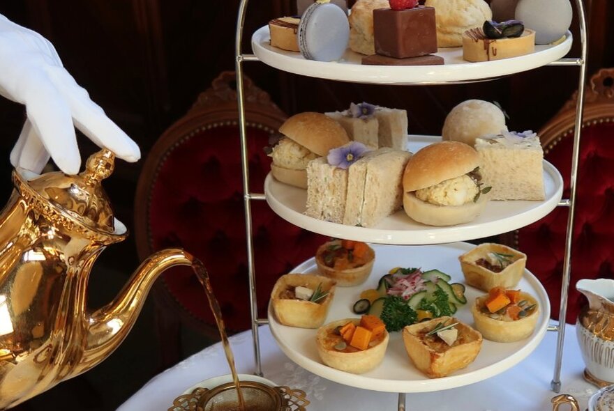 Gloved hands pouring tea from a golden teapot next to a tiered afternoon tea cake stand.
