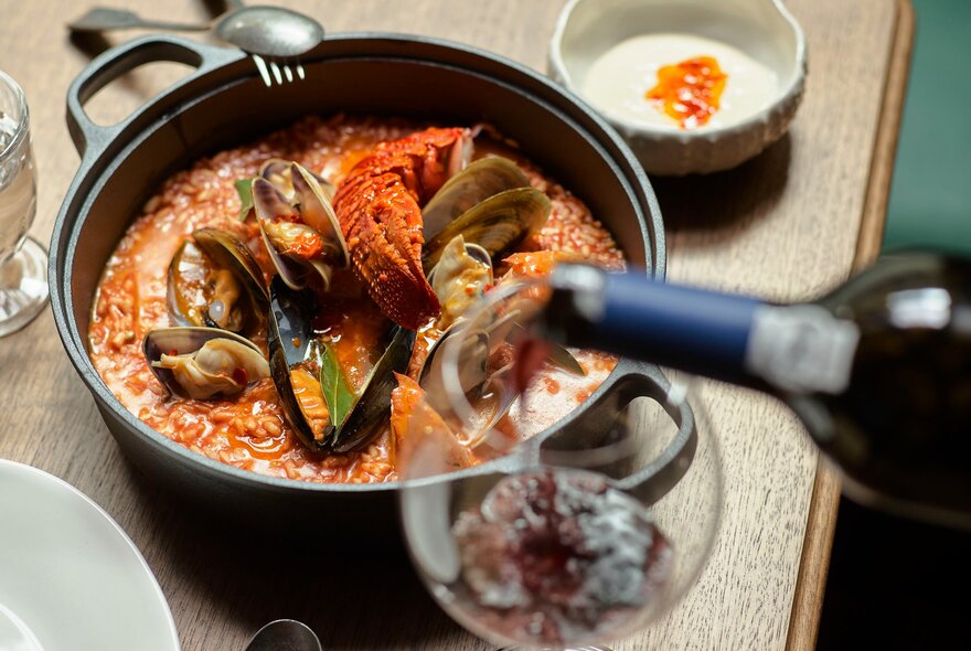 A seafood hot pot with muscles and lobster on a wooden table next to a wine glass in the process of being filled with red wine.