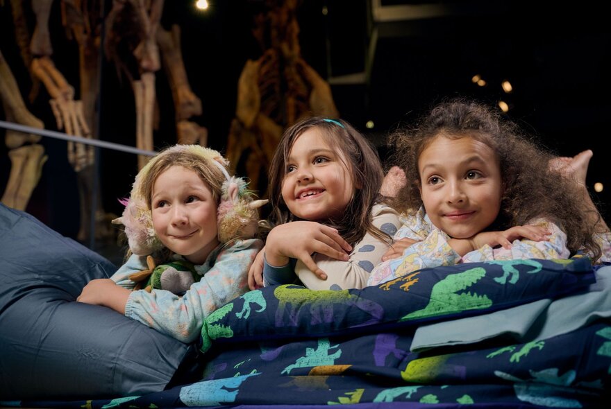 Three smiling young girls lying on a mattress with sleeping bags, inside a museum, next to a dinosaur skeleton.