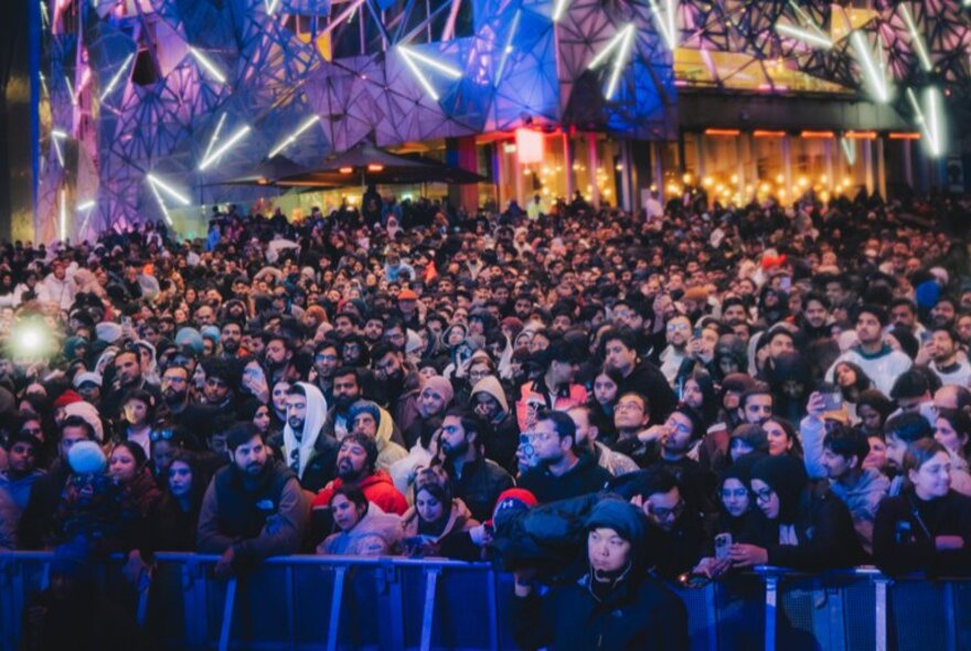 A large crowd of people gathered in Melbourne's Fed Square for an evening event, standing behind a barricade.