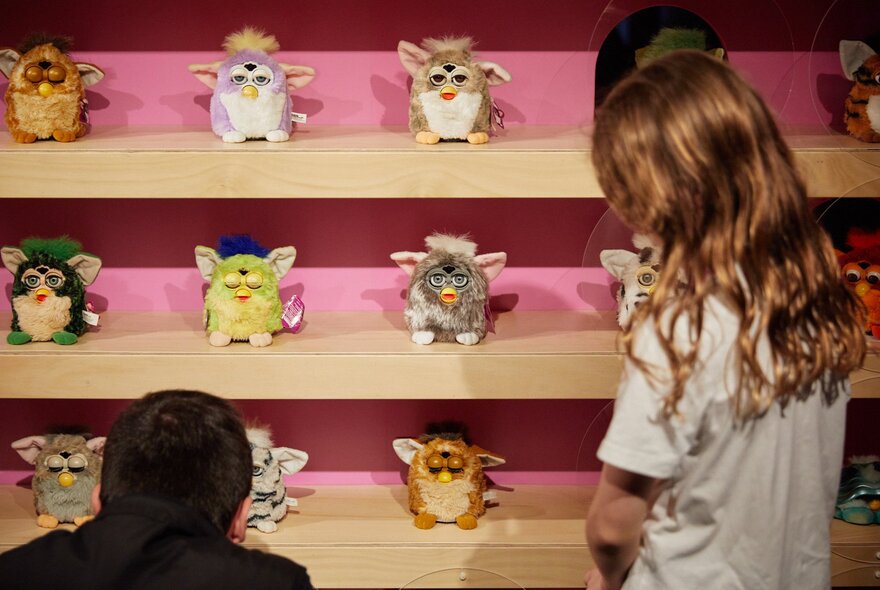 A collection of vintage Furby electronic robotic toys displayed on wooden shelves with two children looking at them.