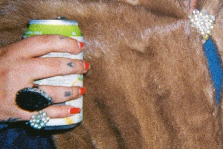 A woman's hand with large rings, tattoos and red nails holding an alcoholic drink in a can in front of a pelt of animal fur.