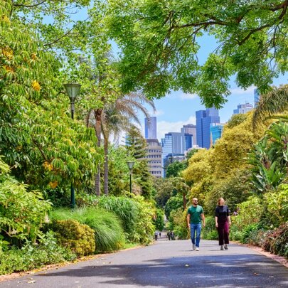 People walking along a plant-lined pathway in public gardens, with city towers in the background.