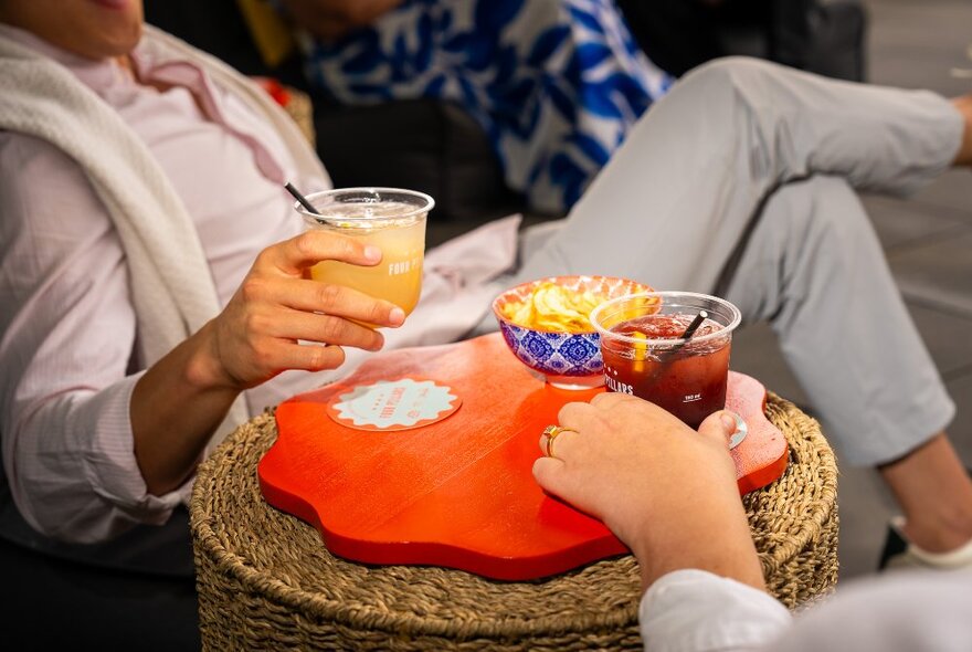 A person in a pink shirt lounging back on a low chair, picking up a cocktail in a short plastic glass off a low wicker stool with wooden platter.