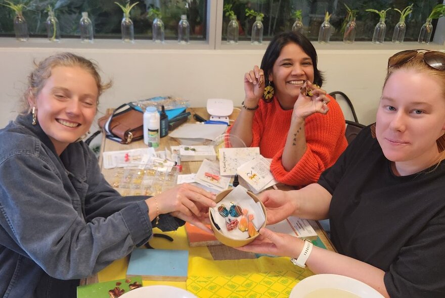 Three workshop participants seated at a table with craft materials, holding containers with jewellery.