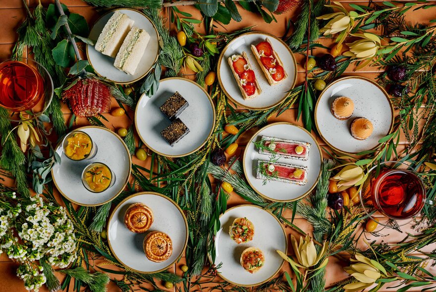 Looking down at eight small plates that each contain two small items of food from a high tea selection, on a table decorated with native flowers.