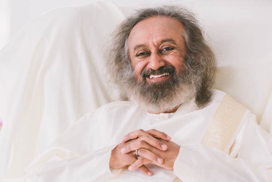 A smiling portrait of meditation guru, Gurudev Sri Sri Ravi Shankar with hands clasped, dressed in white, leaning against a white fabric background.