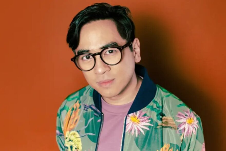 Portrait of comedian, Michael Hing, wearing a floral bomber jacket in front of an orange background.