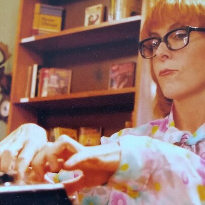 Australian musician and producer Nicole Skeltys, a woman with glasses and red hair wearing a floral shirt fiddles with electronic controls in front of a bookshelf.