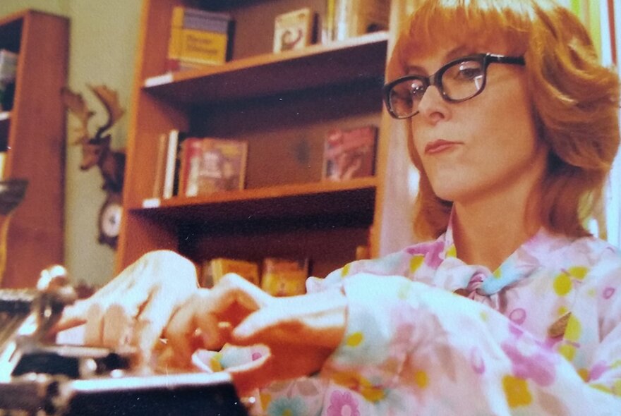 Australian musician and producer Nicole Skeltys, a woman with glasses and red hair wearing a floral shirt fiddles with electronic controls in front of a bookshelf.
