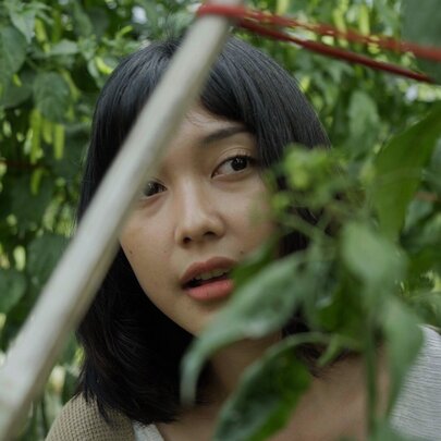 Film still of a young woman glimpsed amongst foliage.
